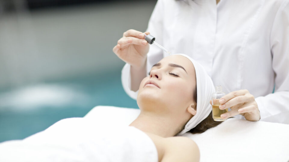 A woman lies on a spa table with her eyes closed while a professional applies facial serum using a dropper.