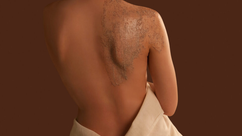 A woman with a towel wrapped around her waist sits with her back exposed, applying a brown body scrub to her shoulder, with a small bowl placed beside her.