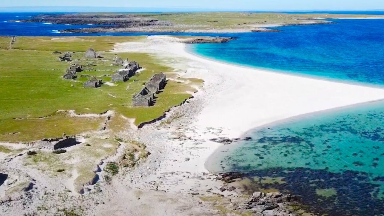 Aerial view of a white sand beach with turquoise water, rocky ruins, green grass, and distant land under a clear blue sky.